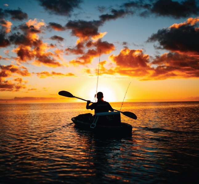 A person kayaking on calm waters during a sunset, holding a paddle and carrying fishing rods. The sky is illuminated with warm orange and yellow hues, contrasting against the dark silhouette of the kayaker and scattered clouds.